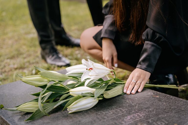 Bouquet de lírios brancos sobre uma lápide, com uma pessoa ajoelhada ao lado em gesto de homenagem.