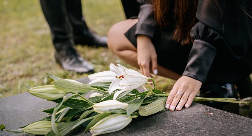 Bouquet de lírios brancos sobre uma lápide, com uma pessoa ajoelhada ao lado em gesto de homenagem.